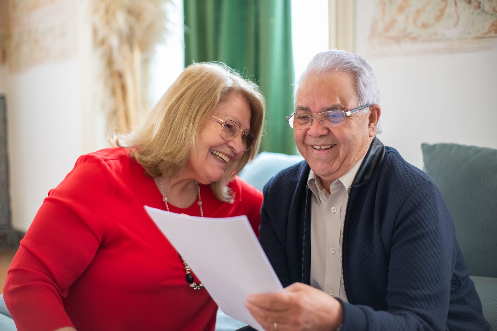 Elderly couple joyfully reading a paper together on the couch in a cozy indoor setting.